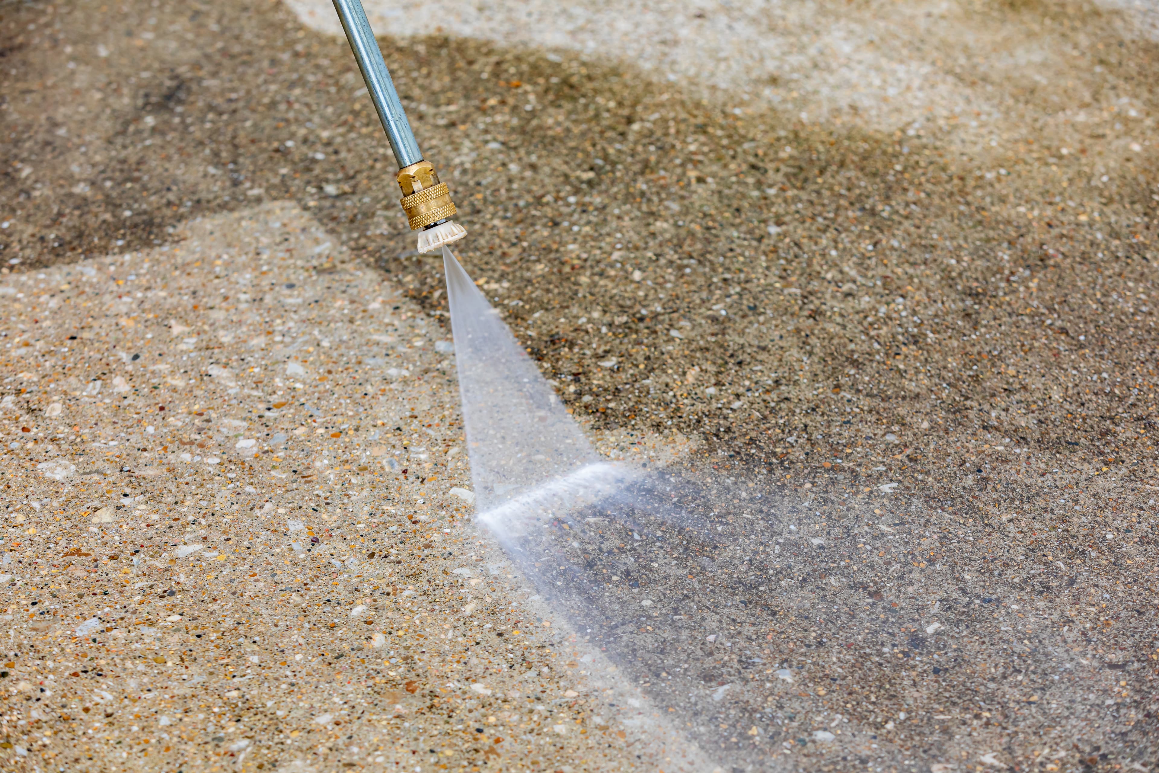 A power washer wand sprays water onto dirty concrete, revealing a clean surface underneath.